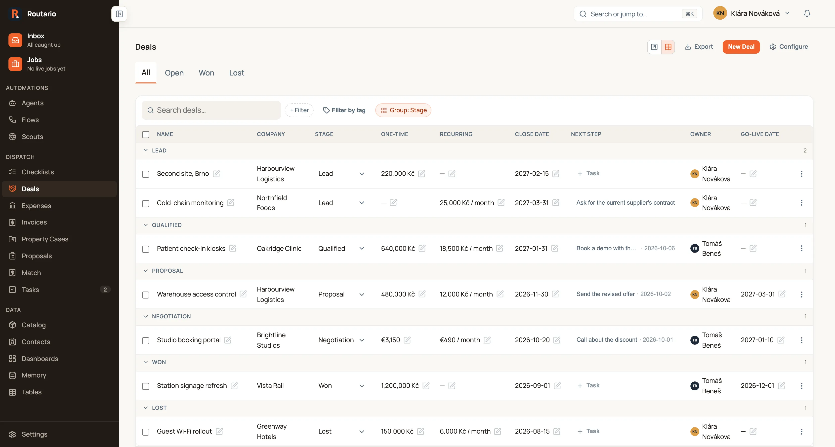 The Deals table, grouped by stage. The toolbar shows Search deals…, + Filter, Filter by tag and a highlighted Group: Stage chip. Headings LEAD, QUALIFIED, PROPOSAL, NEGOTIATION, WON and LOST divide the rows. Columns: Name, Company, Stage (a dropdown on each row), One-time, Recurring, Close date, Next step, Owner and Go-live date. Some deals have both amounts (480,000 Kč and 12,000 Kč / month), some only one, with — for the other; one deal is priced in euros (€3,150 and €490 / month). Next step shows tasks such as "Send the revised offer · 2026-10-02"; rows with no open task show "+ Task".